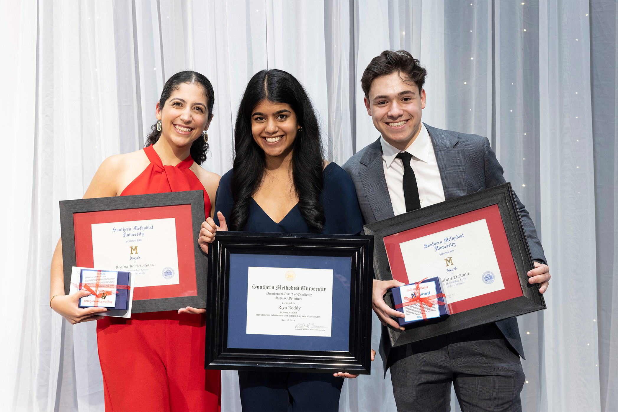 three smiling students holding framed certificates in front of white shear curtain on stage