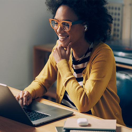woman with glasses in yellow sweater working on a laptop and smiling