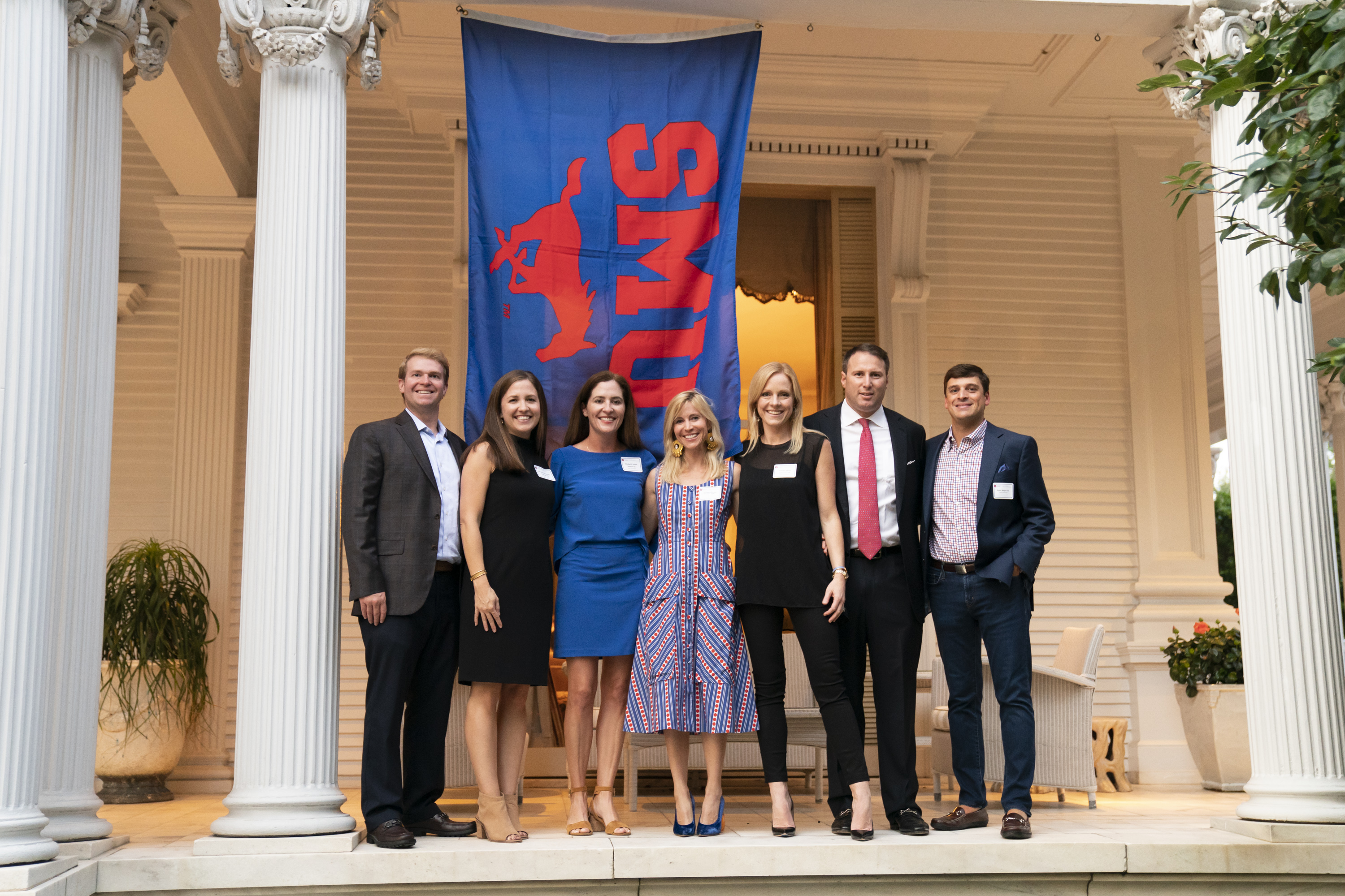 Guests stand outside of the Wedding Cake House in front of an SMU flag.