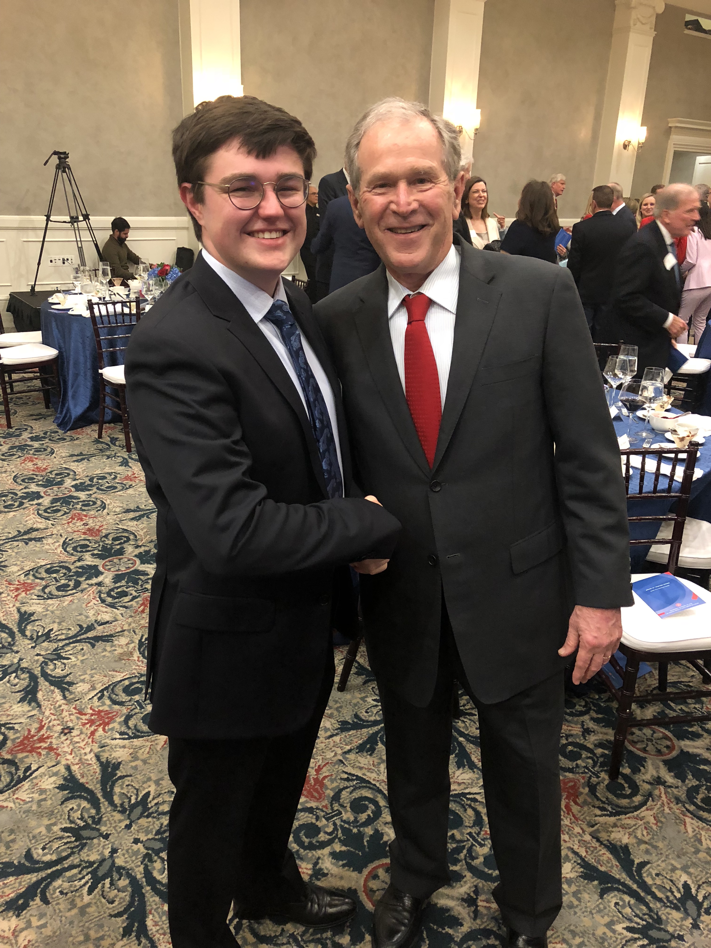 Bradley Potts is a white man with short dark brown hair. He is wearing a black business suit with a navy blue tie. He is shaking the hand of former President George W. Bush, Jr.
