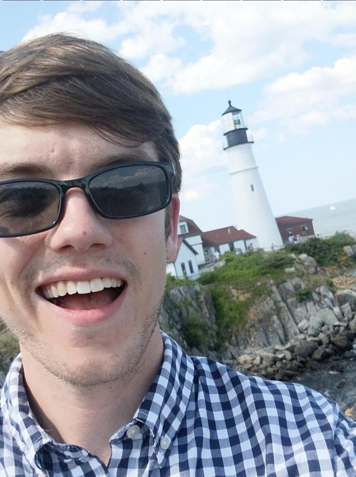Carter Koehler is a white man with brown hair. He wear sunglasses and a white and navy blue plaid shirt. In the background behind him is a white lighthouse.