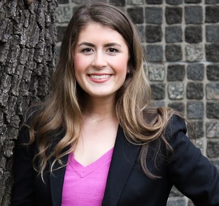 Katherine Boomer is a white woman with dark blonde hair past her shoulders. She wears a pink blouse and black blazer and stands in front of a tree.