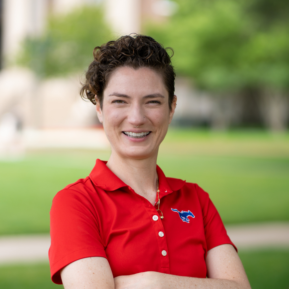 Alexandra Thibeaux is a white woman with short curly hair, cut in a pixie style. She is wearing a red short-sleeved polo with a blue mustang embroidered on the left pocket area. The background is the blurred path towards Dallas Hall.