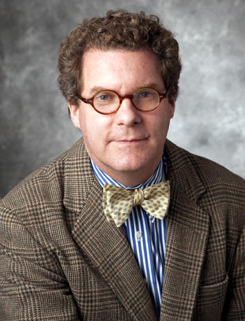 Dr. David D. Doyle is a white man with brown curly hair and round tortoise shell colored glasses. He wears a brown blazer, a gold bow-tie, and a light blue button up oxford shirt.