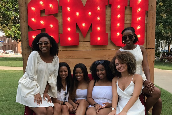 6 female black students in white dresses sit on a red couch in front of a large marquee sign for SMU - photo taken during a Boulevarding event.