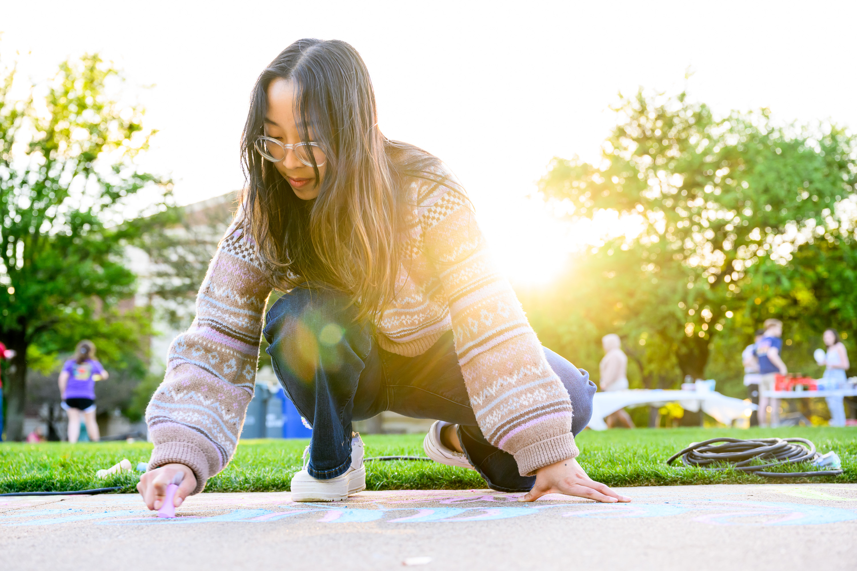student drawing on sidewalk with chalk outside with sun shining and green grass and trees in the background