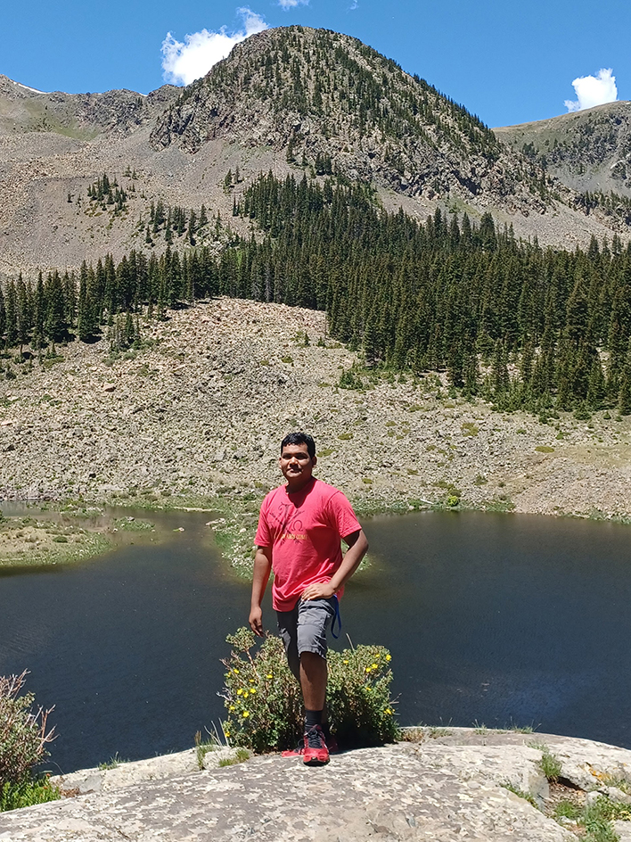 Taos student Giancarlos Dominguez stands on a rock with a mountain lake behind him.