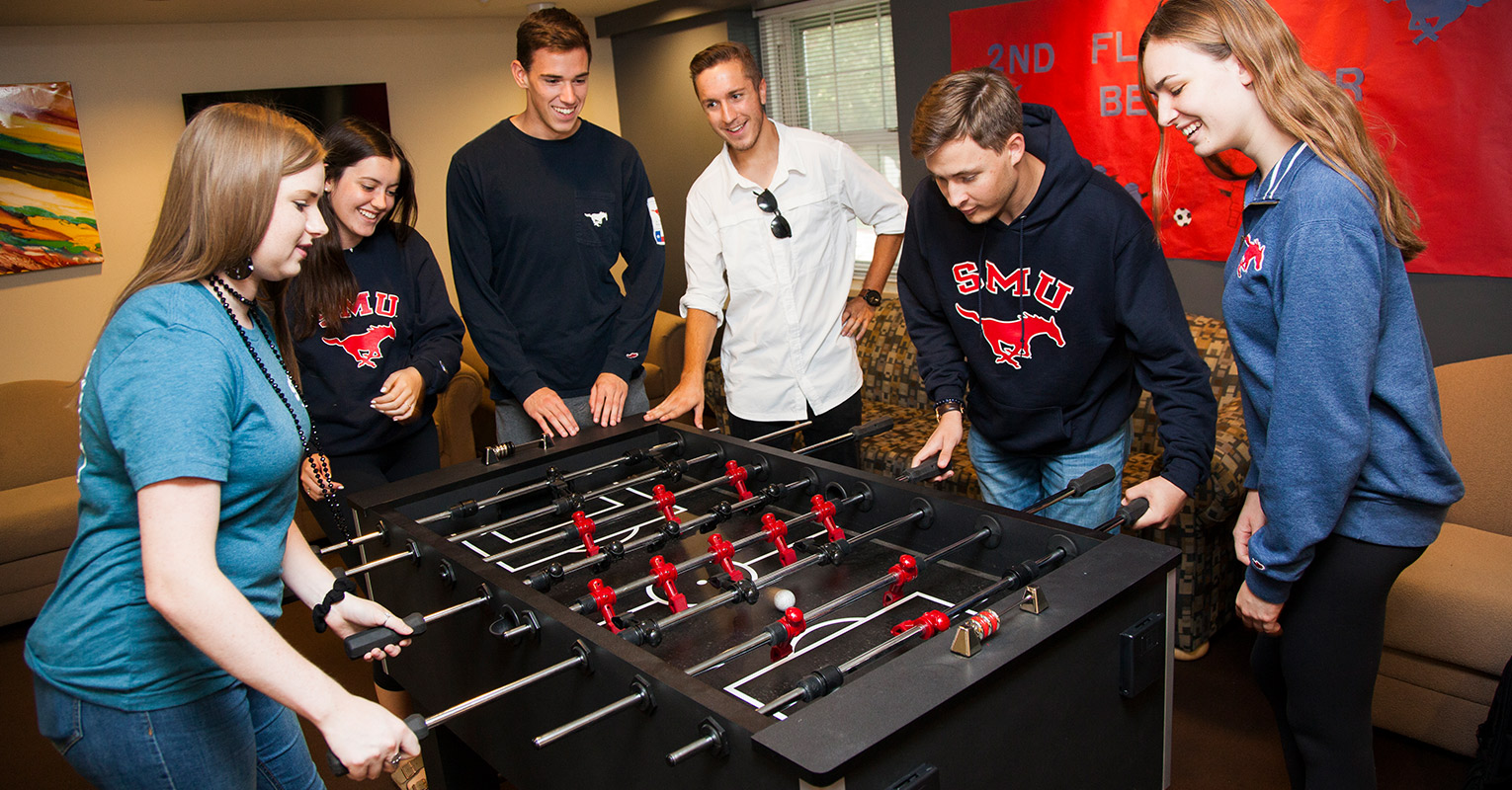 A crowd of people gathered around a foosball table while two players compete against eachother. 