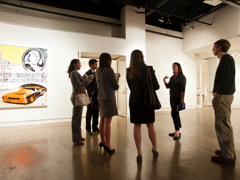 A group of online MBA students wearing professional attire stand in a circle listening to a host at an art gallery