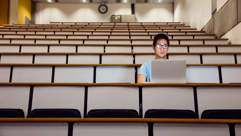 A man with glasses sits in an empty lecture hall while working on his computer