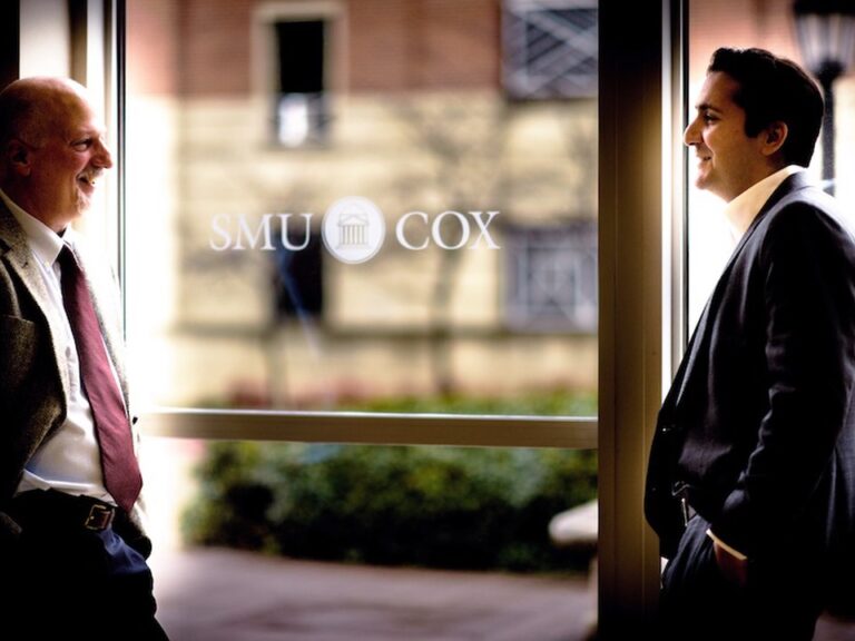 Two SMU Cox faculty members have a conversation in front of a window that has the school emblem printed in white