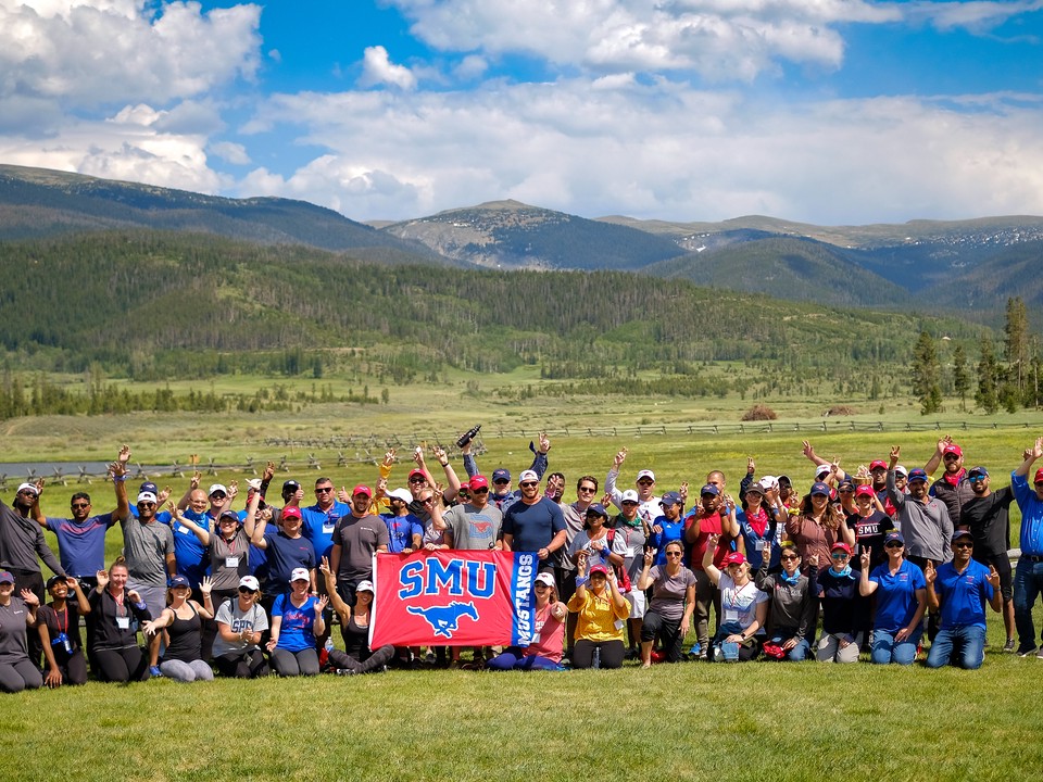 A group of SMU Cox Online MBA students pose for a photo in front of the mountains while holding a red SMU banner