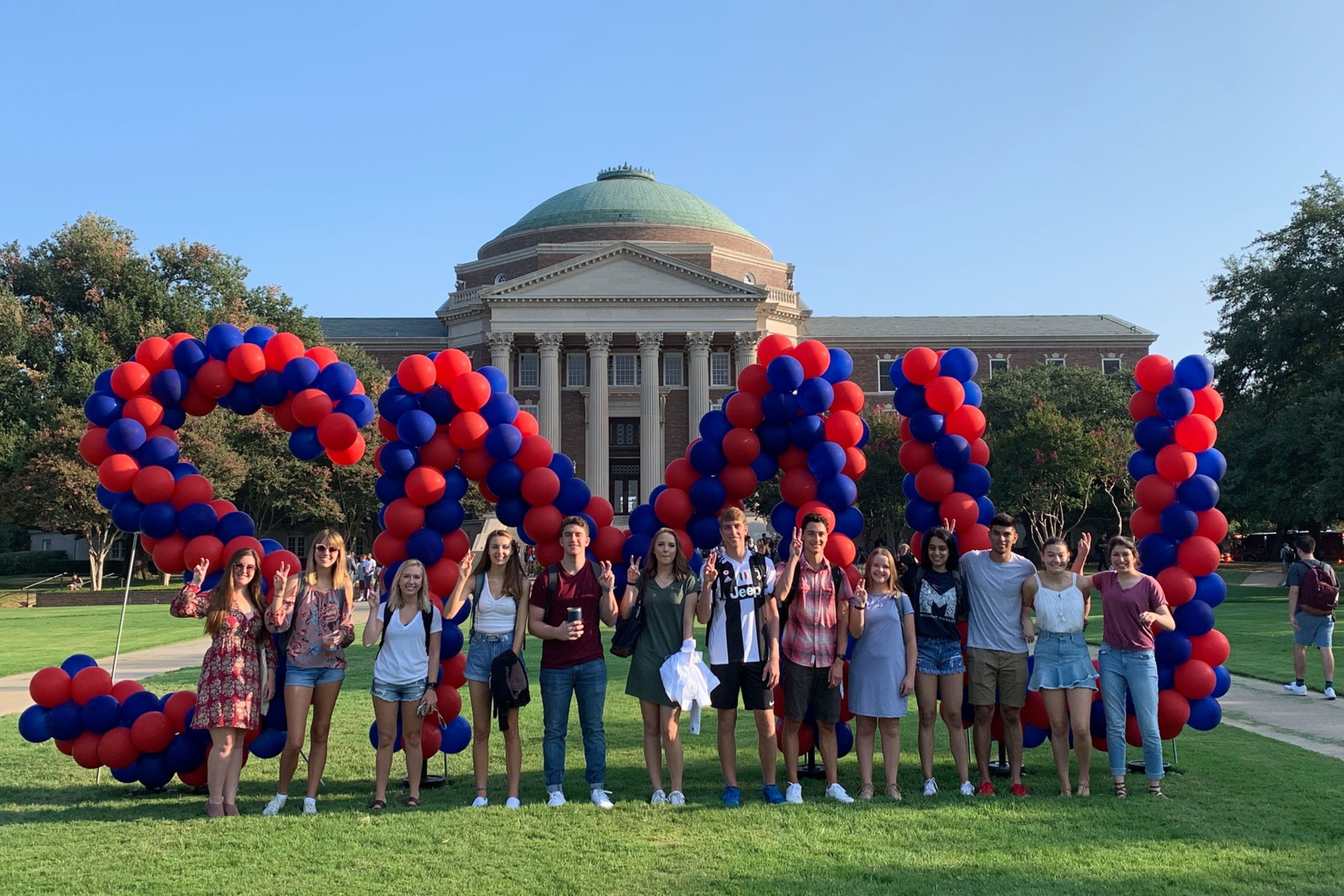 Students stand on Dallas Hall Lawn in front of red and blue SMU balloons.