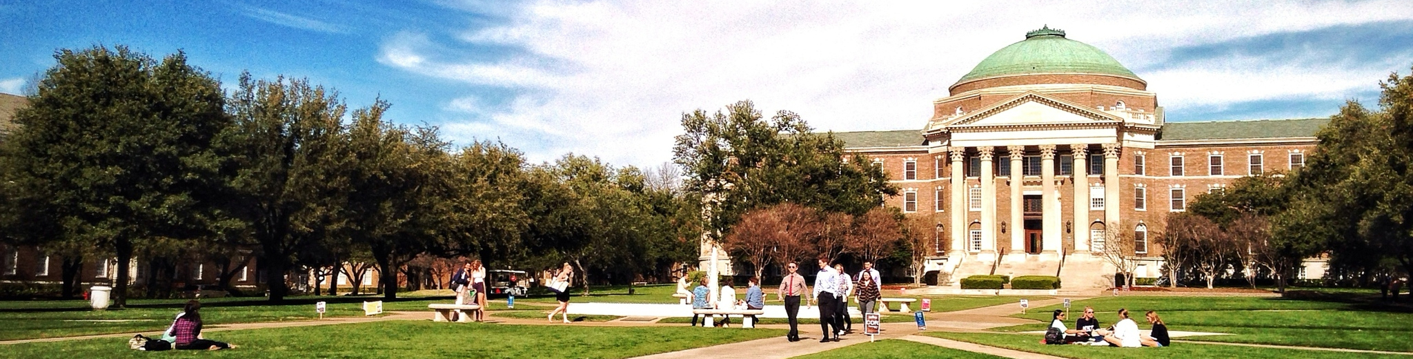 The front lawn of Dallas Hall featuring groups of people walking across the quad and sitting in the grass.