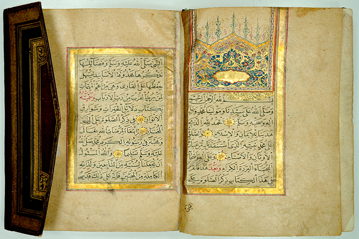 An open Islamic prayer book, introduced by a gilded decorative panel with an arch-shaped ornament above. Part of the pointed envelope flap binding is visible.