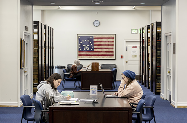 two students study at a large table with book stacks behind them. A flag hangs on the wall.