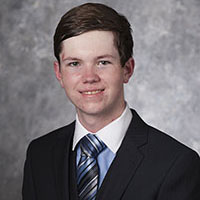 Jared Burleson is a white man with brown hair. He is wearing a blue button-up shirt with a white collar and a black blazer. His tie is striped blue, white, and black.