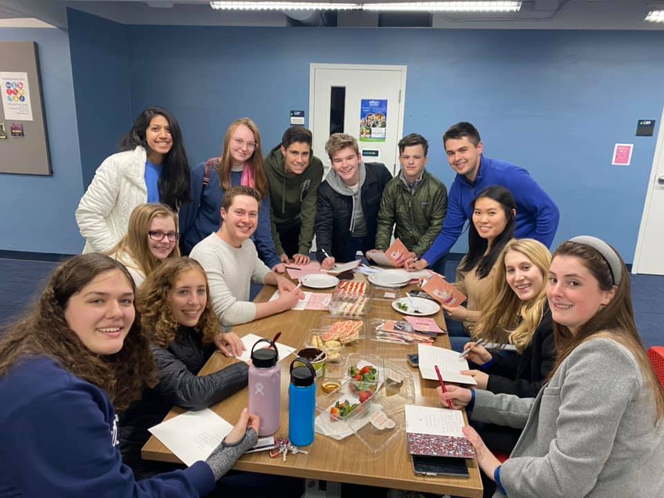 13 Students are gathered around a Scholars' Den study table writing in cards made from construction paper. On the table are Valentines-themed snacks such as cookies and strawberries.