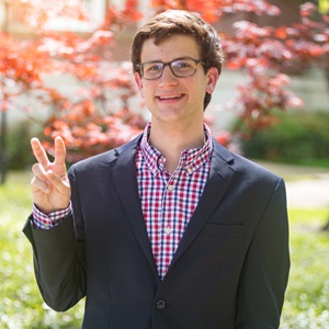 Nicholas Fontela is a white male with short brown hair. He is wearig glasses, a red and blue plaid button up, and a navy blue blazer. He has his hand up in the "Pony Up" hand sign.