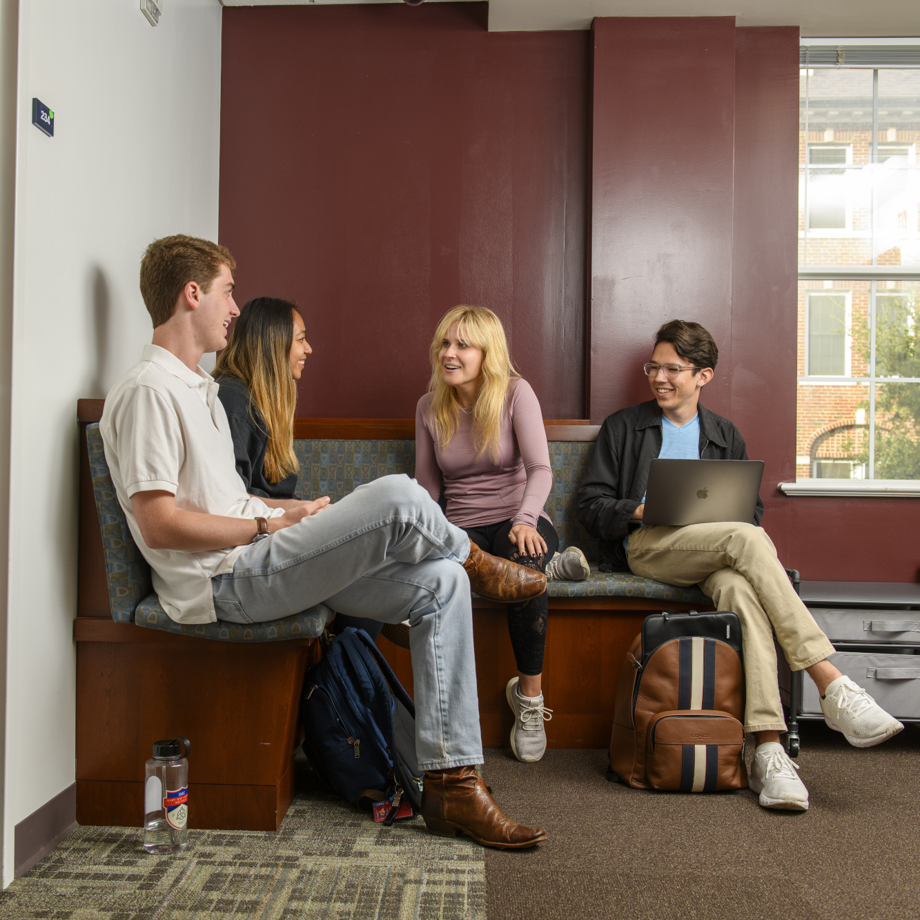 Four students sitting on indoor wooden benches, talking to each other