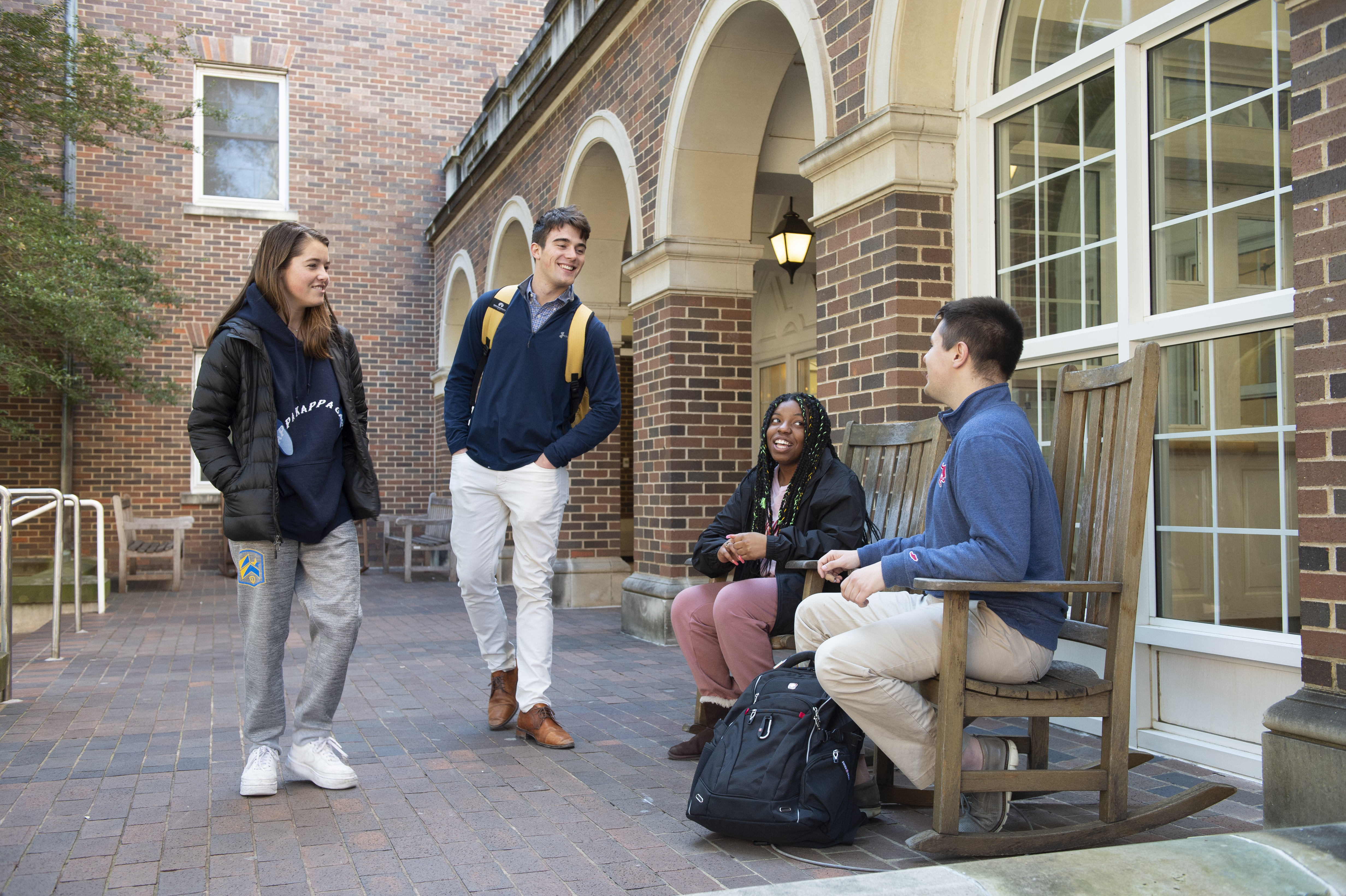 Two students sitting in rocking chairs talk to two students standing up in front of a residence hall.