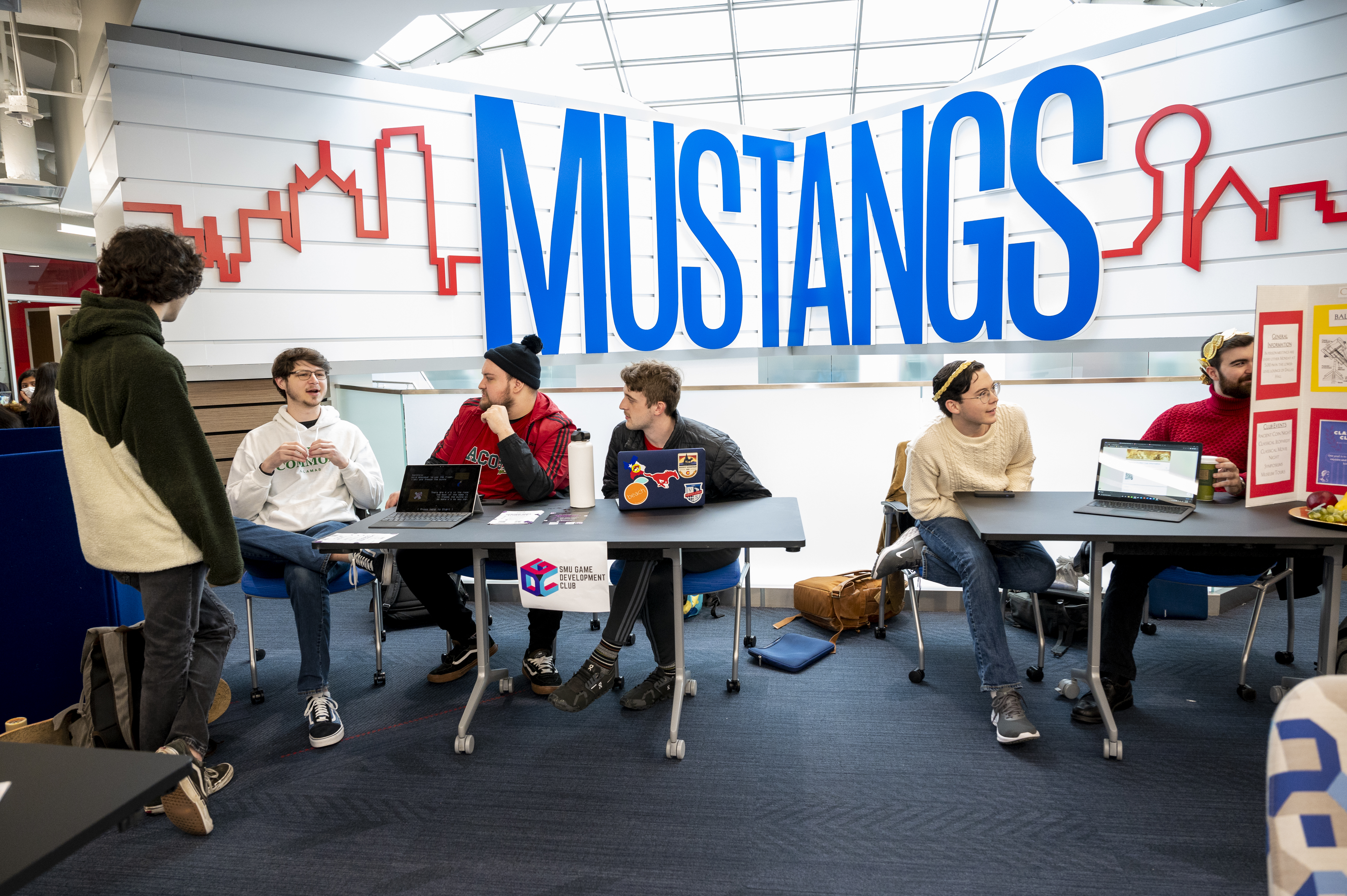 4 students sit around a table and 2 students sit around another table. Large blue letters spelling MUSTANGS along with a red outline of the Dallas skyline is in the background of the tables.