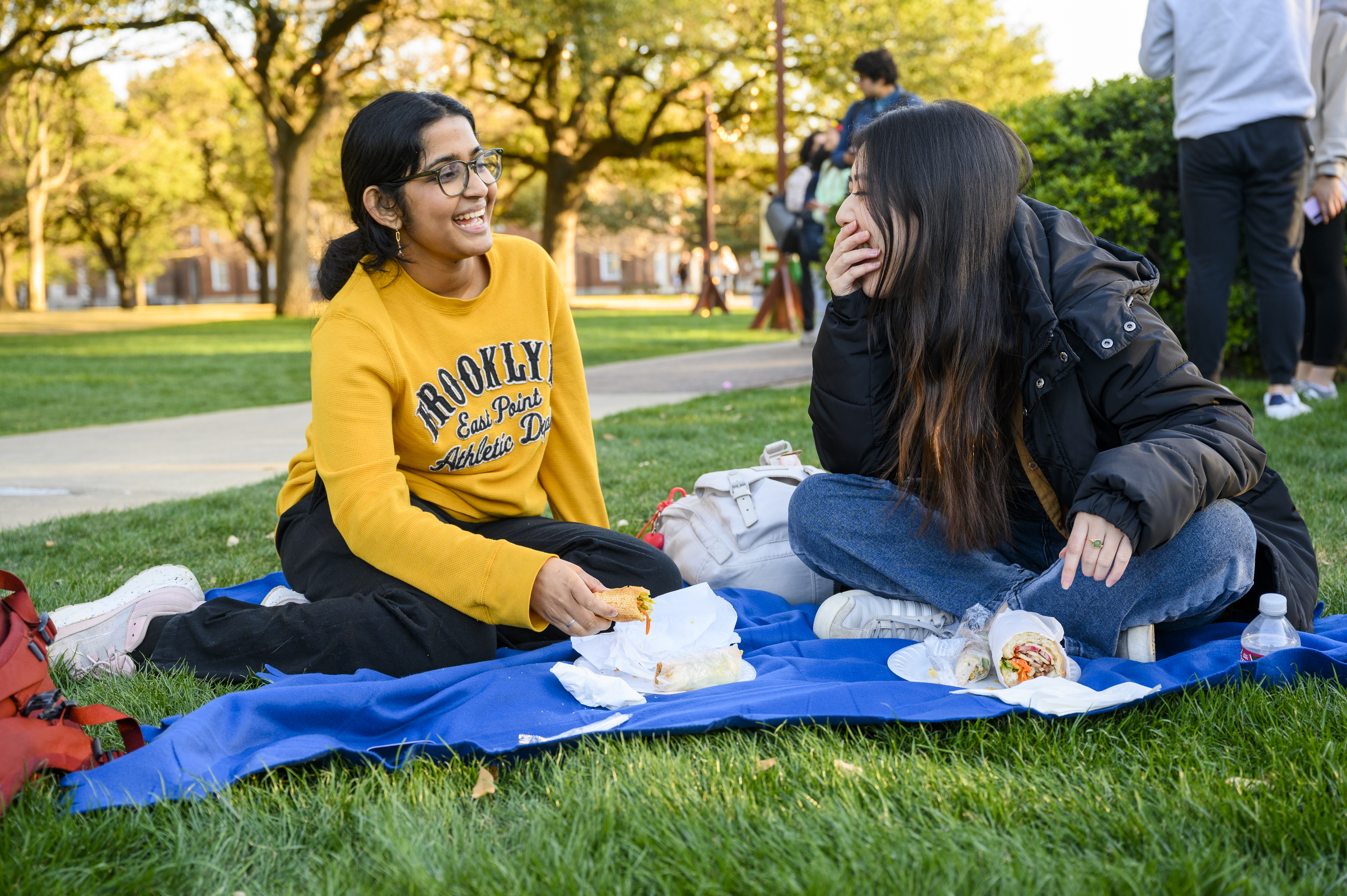 Two students sit, laughing and talking, on a blanket on the lawn of Dallas Hall
