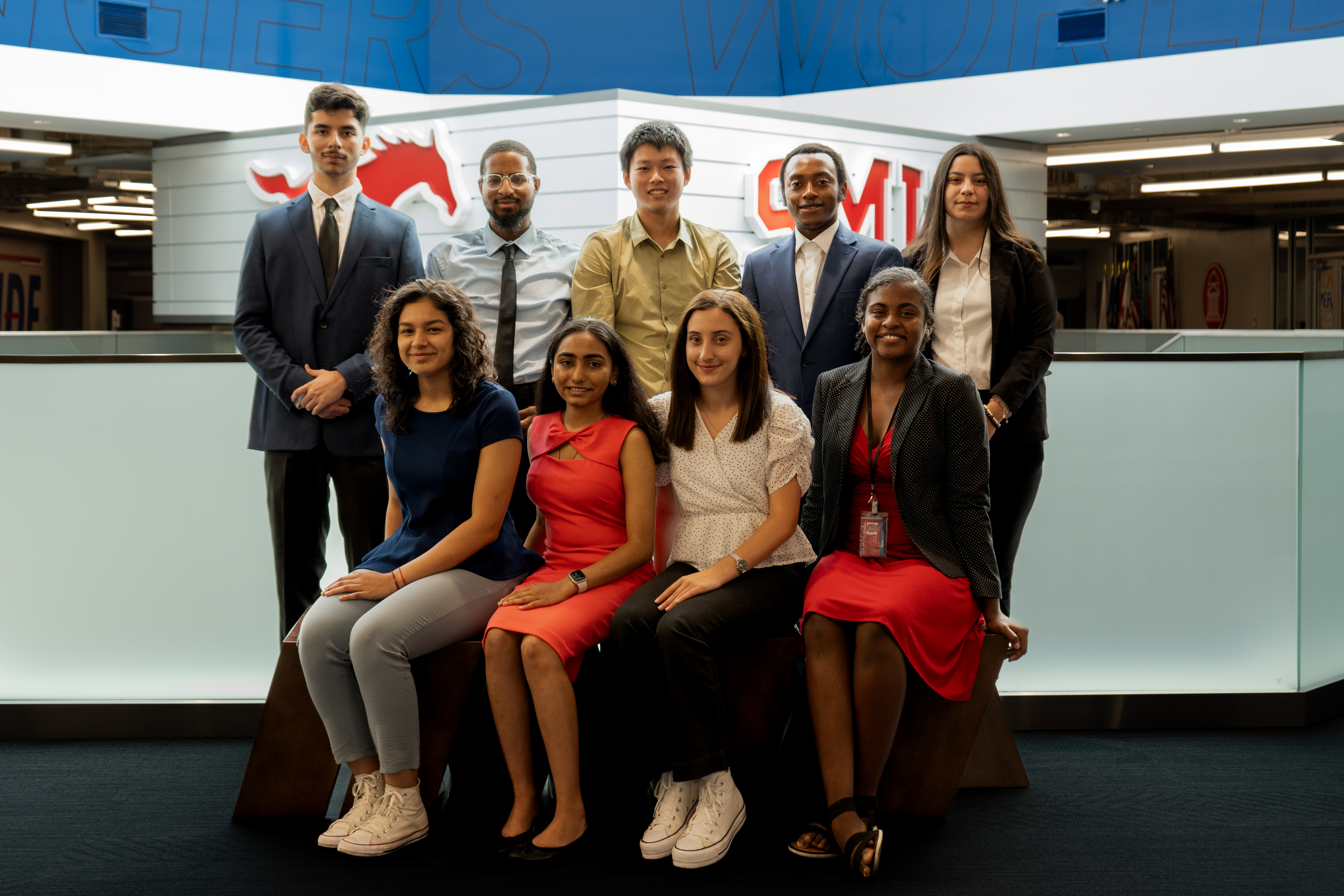 9 Students pose wearing business casual. 5 are standing on the back row, 4 are sitting in front on a bench.