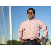 Rahfin Faruk is a Bengali American man with very short black hair. He is wearing glasses, a pink long-sleeve button up, and navy blue slacks. He stands in front of the Margaret Hunt Hill Bridge in Dallas.