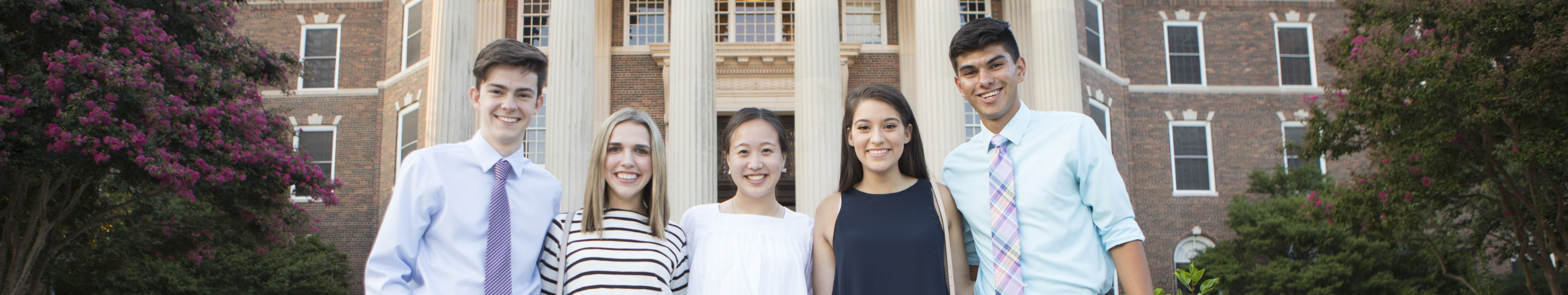 Five students stand with their arms around each other and the iconic pillars of Dallas Hall visible behind them.