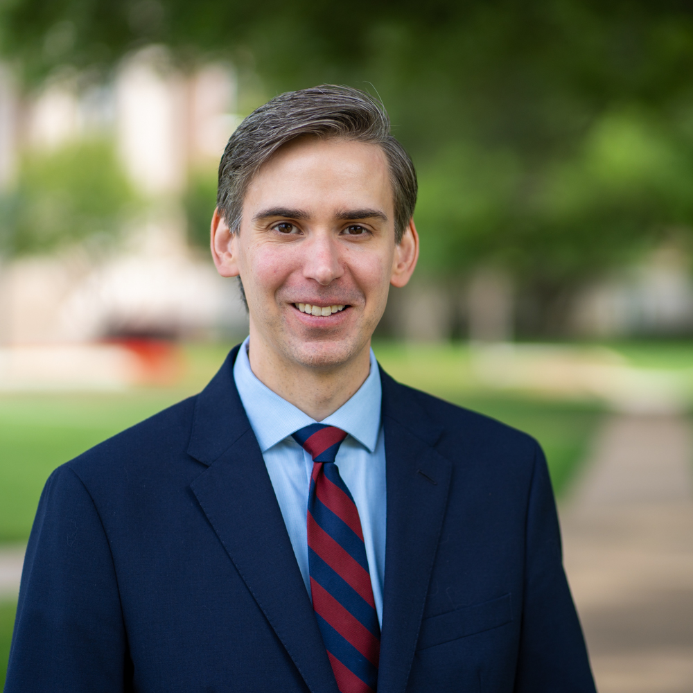 Dr. Brandon Gray Miller is a white man, dressed in a navy blue suit jacket, light blue oxford button up, and red & navy blue striped tie. The background is a path towards Dallas Hall.