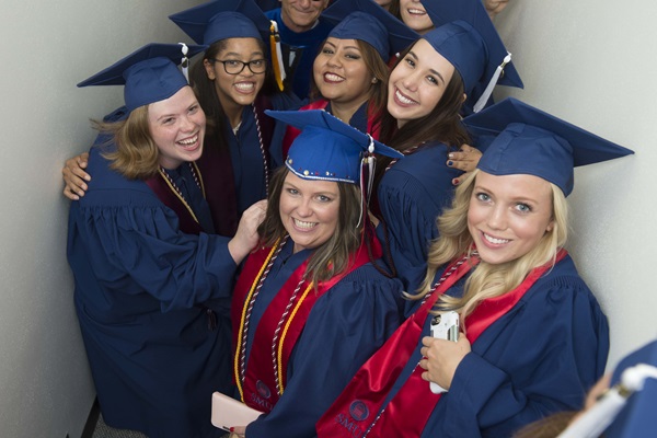 Six students in graduation gowns and honors cords smile at the camera as they take a group picture, viewed from slightly above