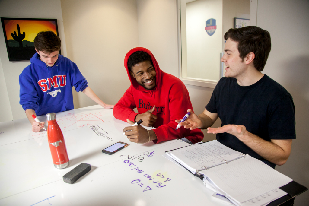 Three students talking as they study at a table. Math notes are written in expo marker on the table in front of them.