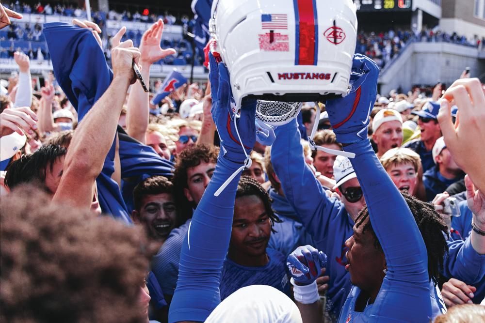 An SMU football player raises a Mustangs helmet amid a cheering crowd.