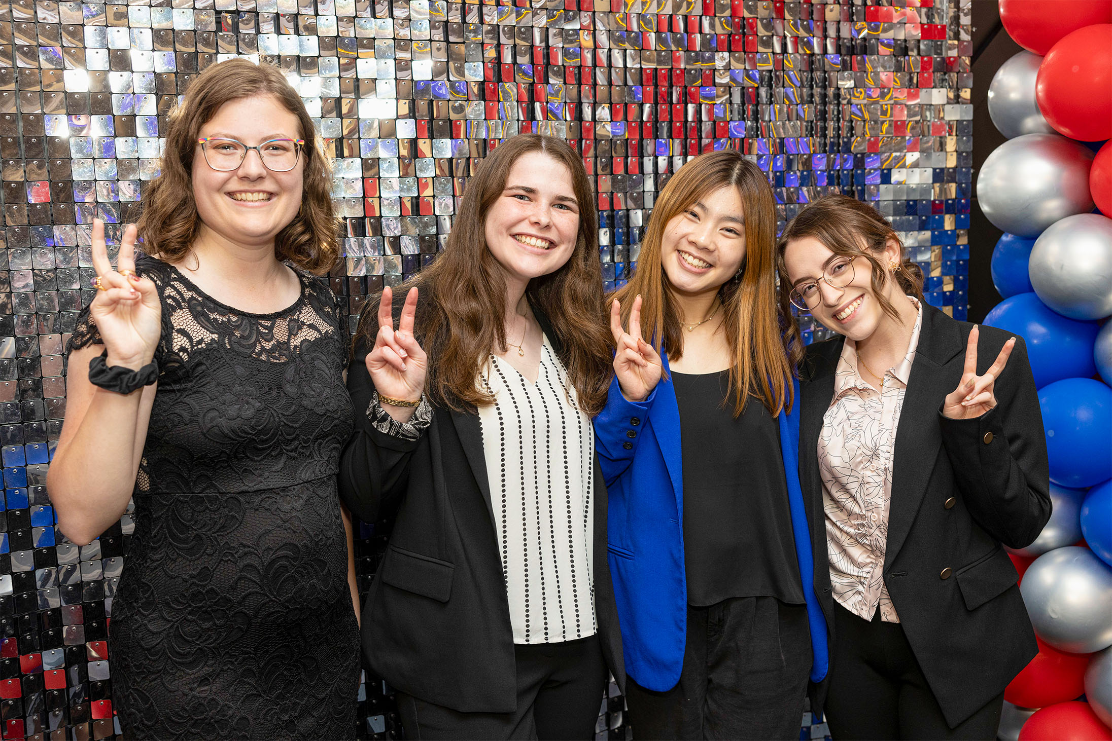 four students with pony up hand signal in front of silver sequined wall with red, blue, and silver balloons
