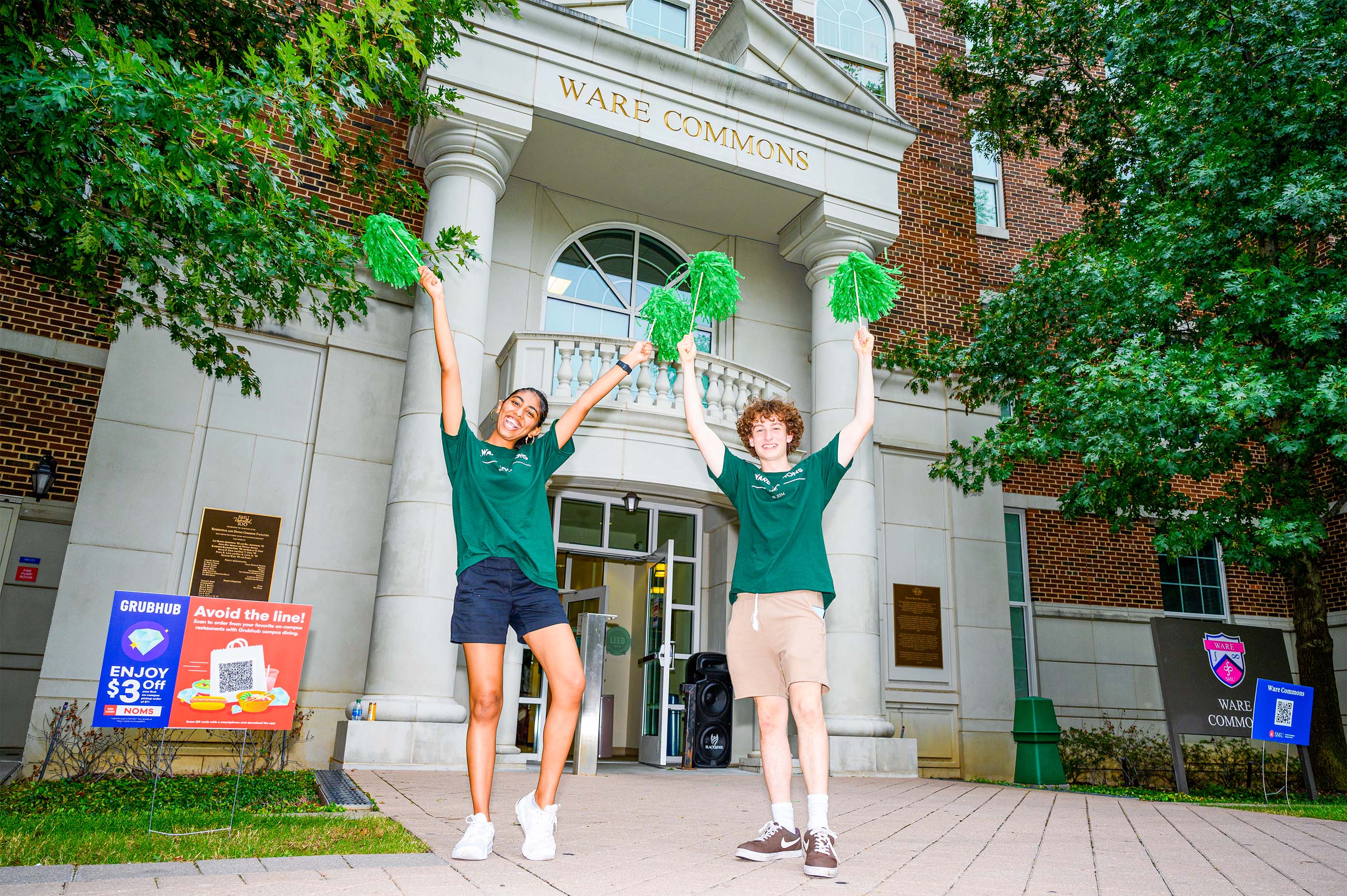 two students excited in front of Ware Commons with green pom poms and wearing green Ware t-shirts