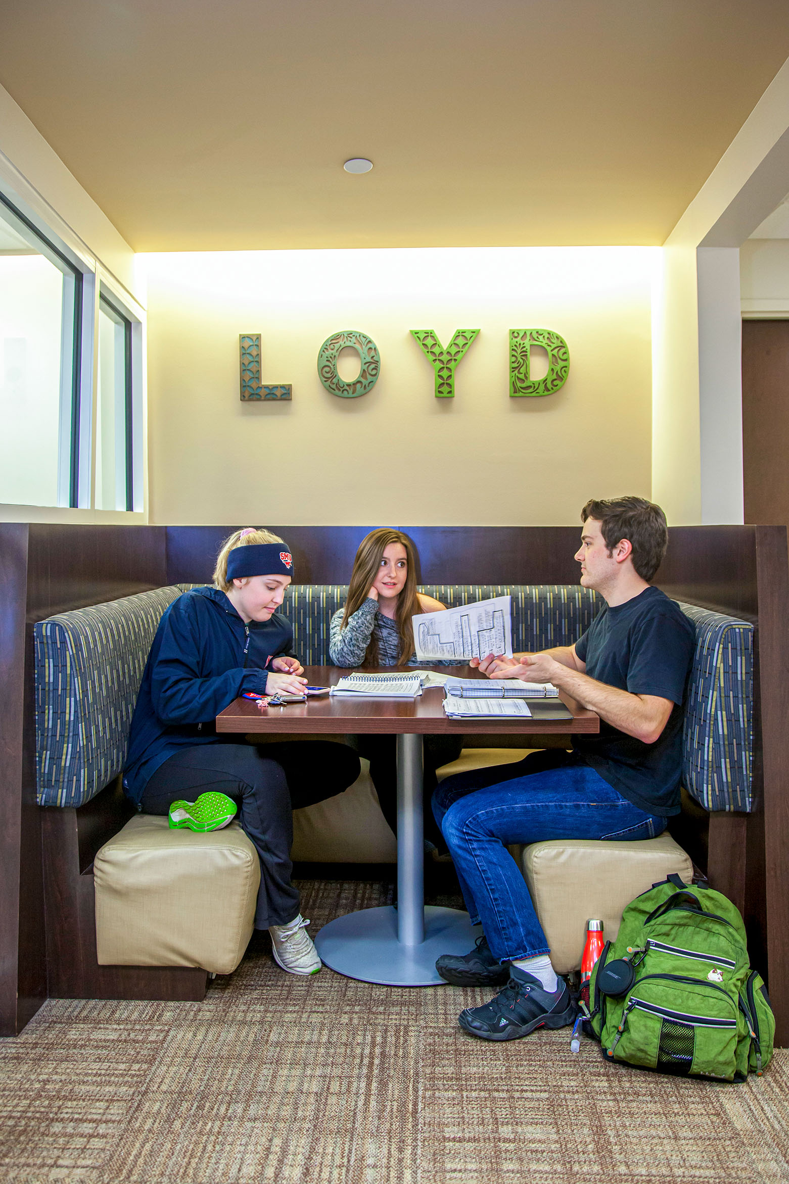 three students sitting at table with books, papers, and a green backpack in loyd commons