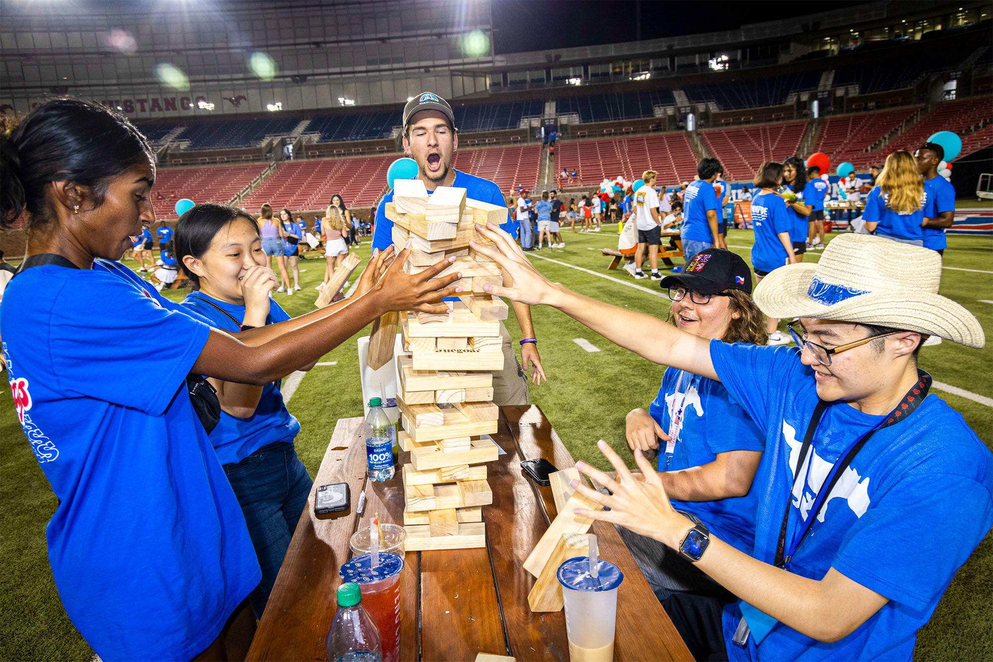 five students in blue shirts playing jenga on picnic table in football stadium at night