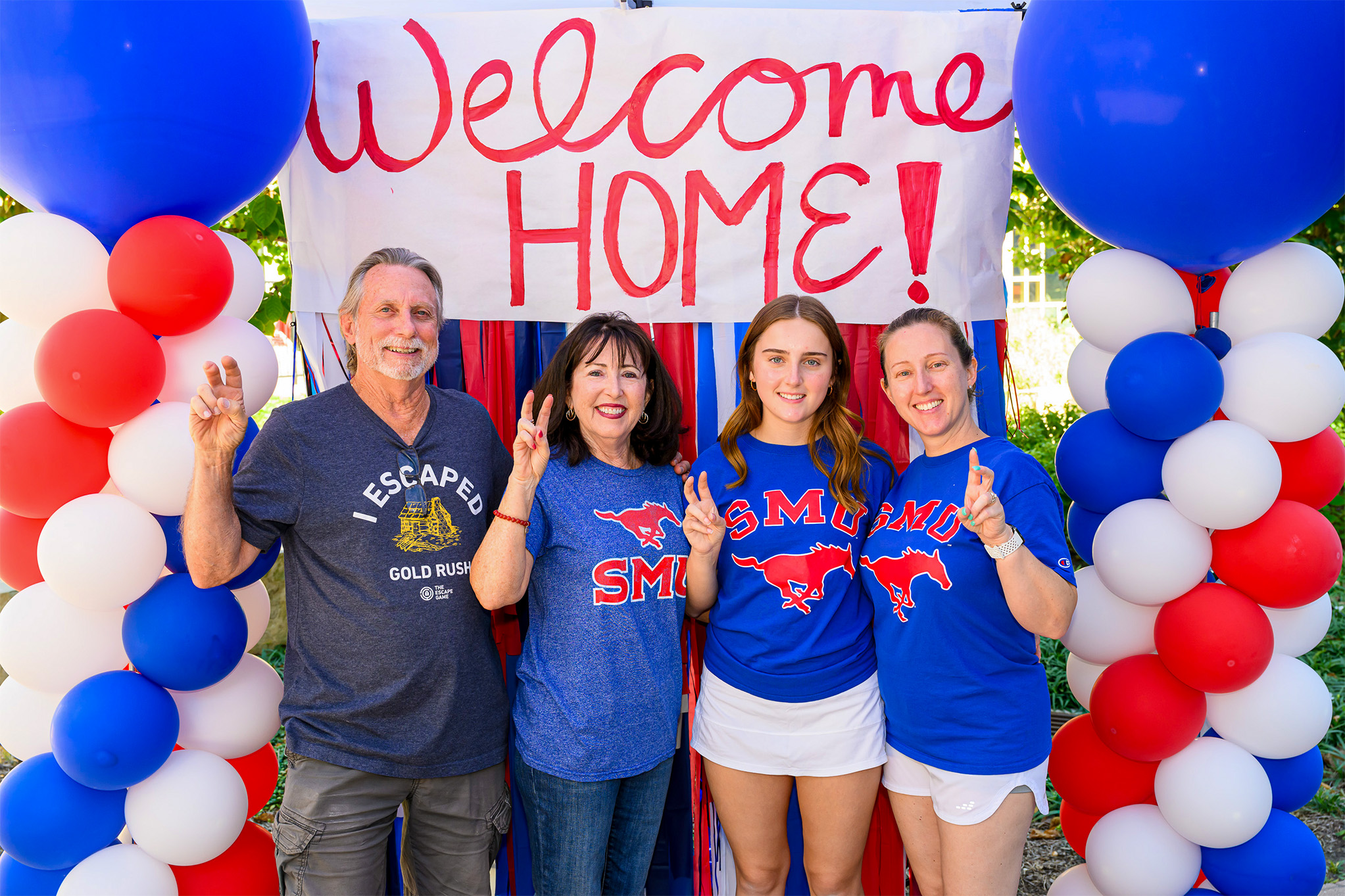 SMU family standing outside holding Pony Up hand sign in front of Welcome Home banner with red, blue, and white balloon towers