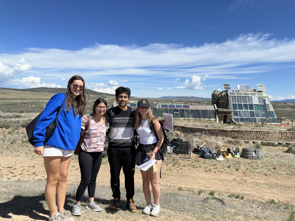four students standing in front of a desert landscape with a earthship house in the background