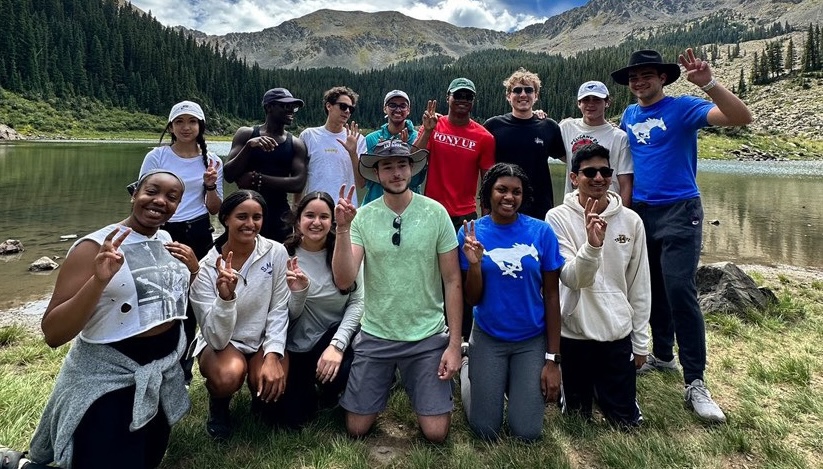 Group of students standing in front of lake smiling at camera doing the Pony Up sign with mountains in the background