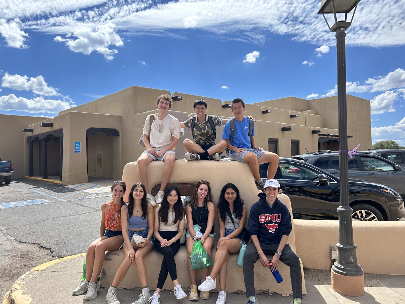 Group of students sitting on a Adobe structure on the sidewalk of a town all smiling at the camera 