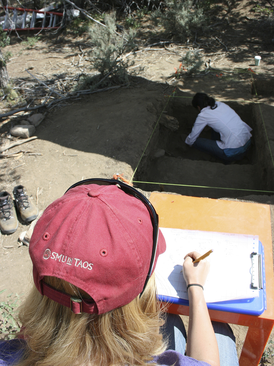 One SMU-in-Taos student takes notes on a clipboard as another digs in a roped off area of the ground.
