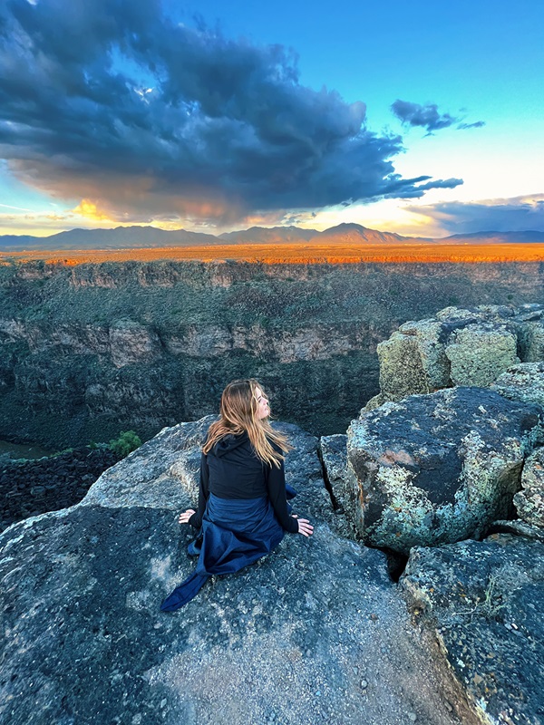 SMU-in-Taos student Jules Campbell sits on giant rocks at the edge of a canyon in front of a Taos, New Mexico sunset. 