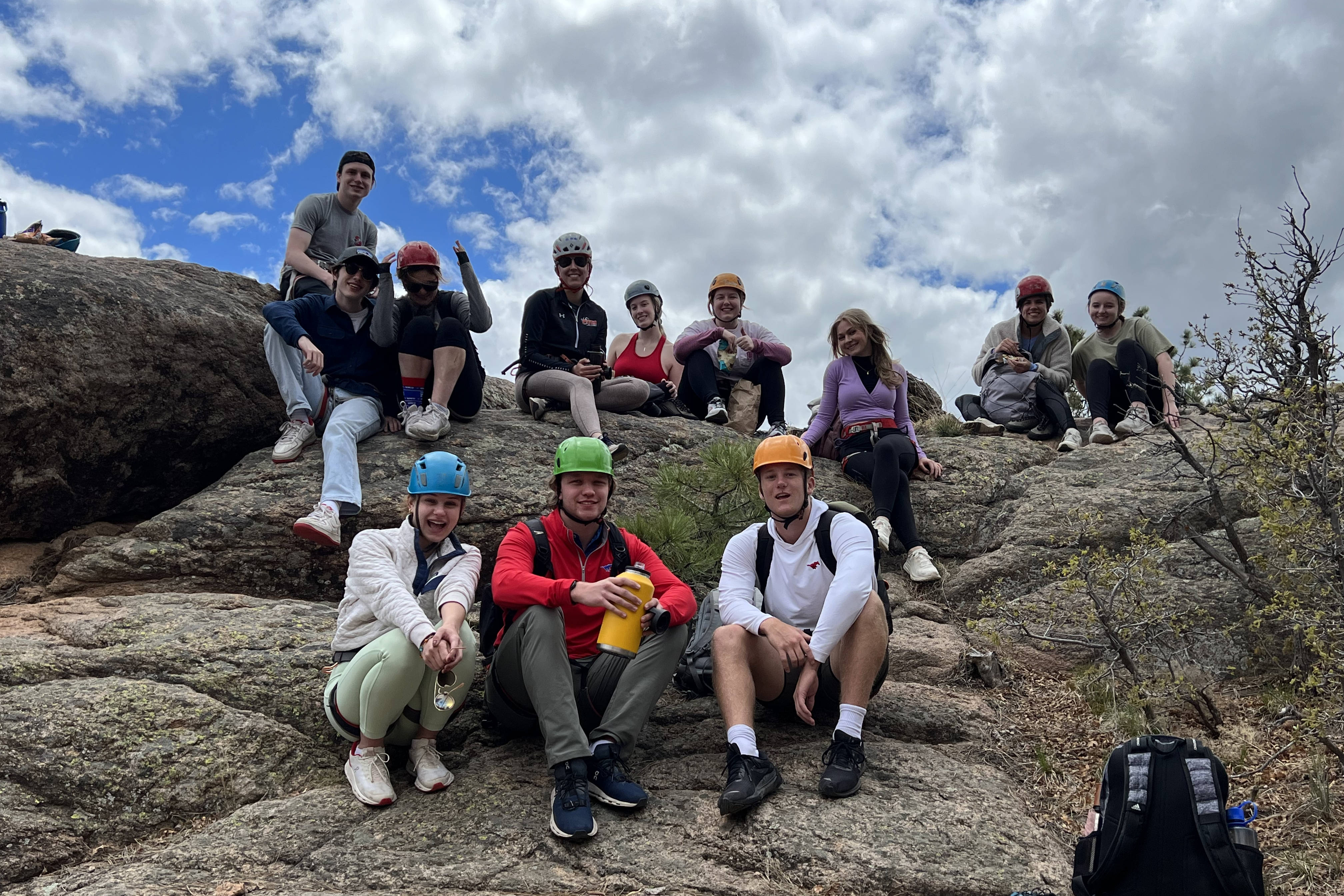 Taos students in rock climbing gear sitting atop a large rock with a blue sky and white clouds in the background.