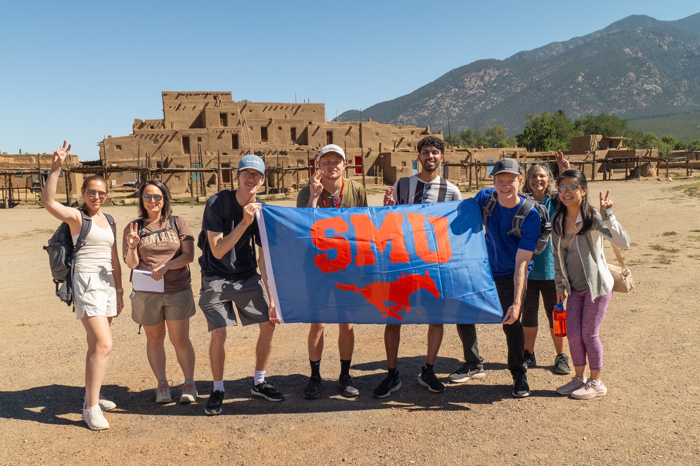 Students standing in front of Taos Pueblo with an SMU flag smiling at the camera and doing the Pony Up hand signal.
