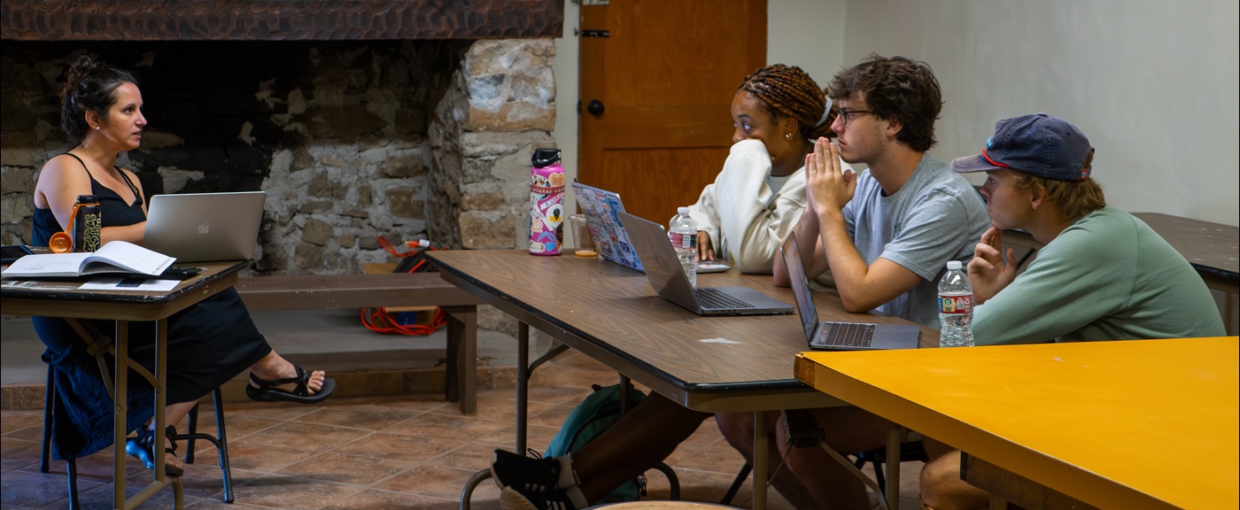 Three students sitting at a table listening to a professor speaking sitting across from them in front of a stone fireplace