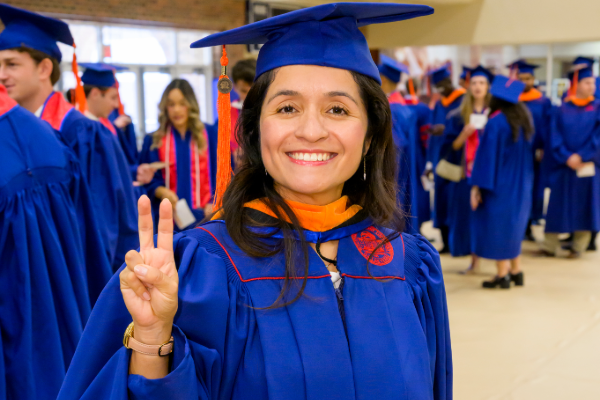 SMU graduate Tania Caldua Cuﬀ stands in her graduation robe giving the SMU pony ears hand signal
