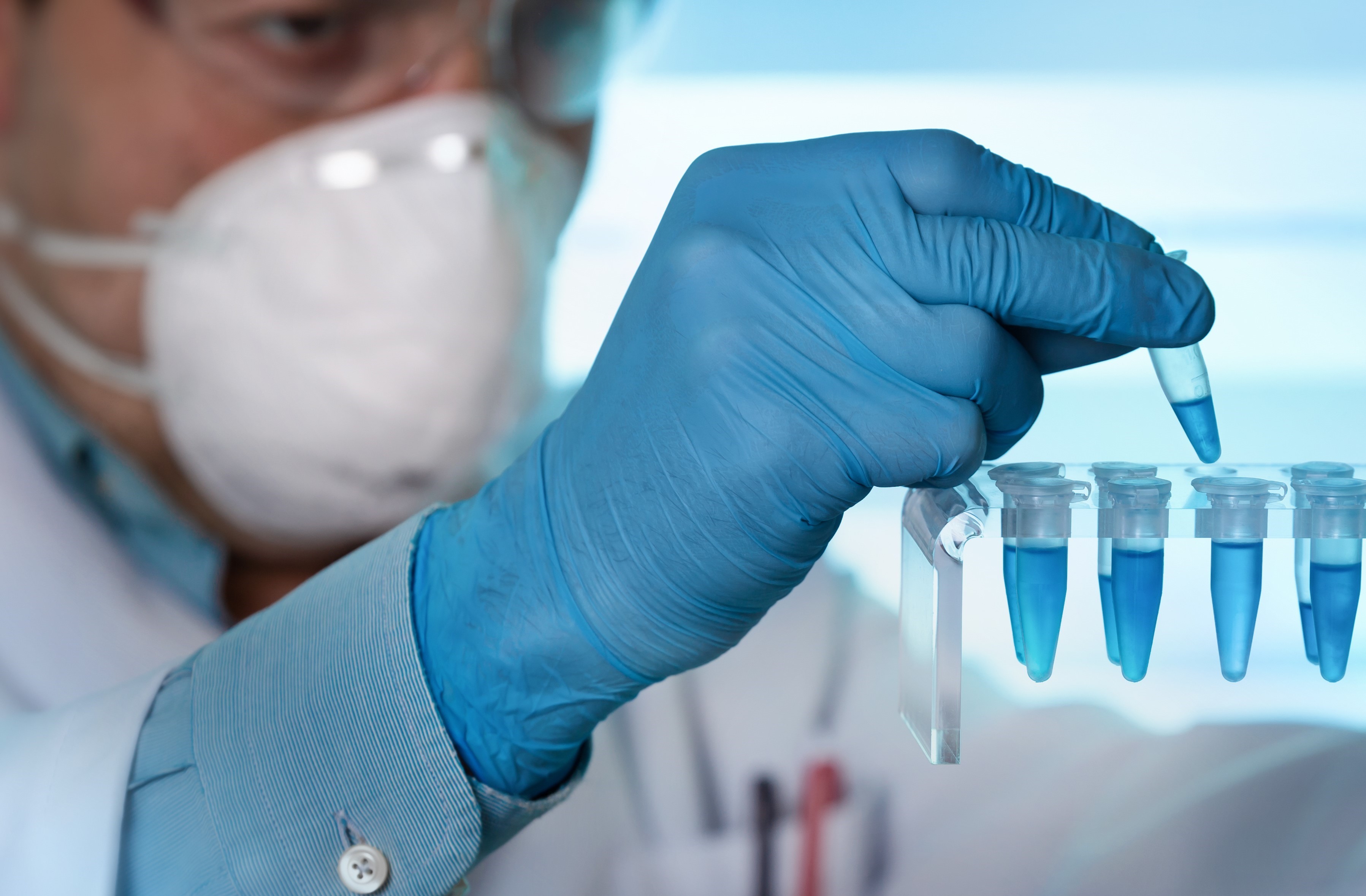 A scientist wearing a mask and blue gloves handling test tubes in a laboratory setting.