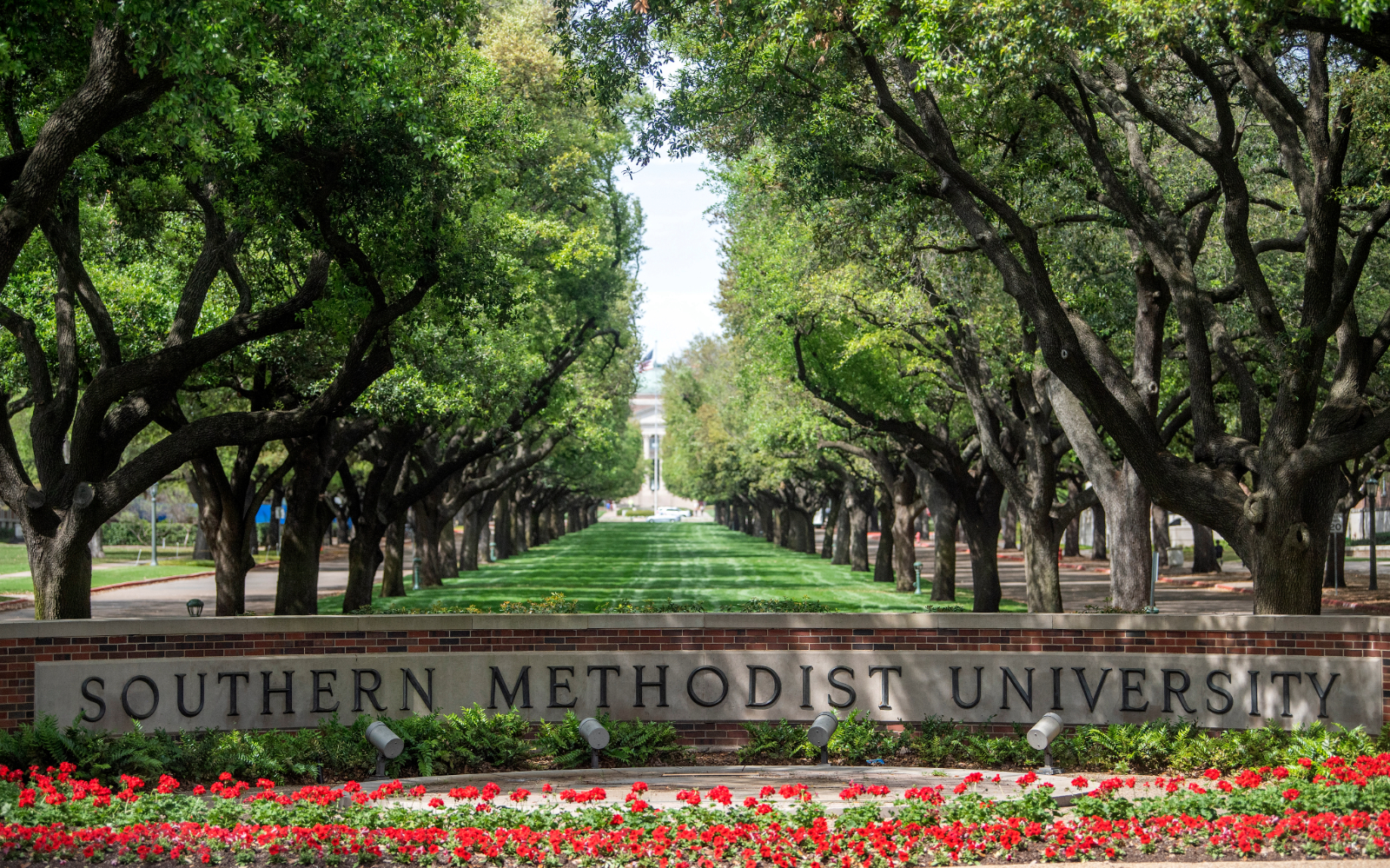 A view of the entrance to SMU with a landscape of tress and flowers and a stone sign reading Southern Methodist University on a lush spring day